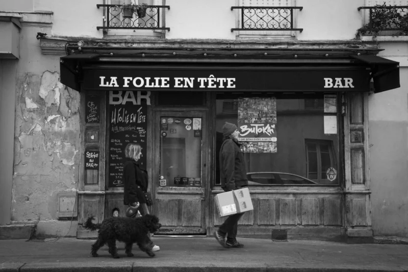 Thierry · 08/03/2024 · 3840 x 2560 Cette photographie en noir et blanc capture une scène urbaine devant un bar nommé **« La Folie en Tête »**. Le sujet principal est un homme tenant un chien en laisse, accompagné d’une femme avec un sac. La composition met en valeur la façade du bar, avec ses enseignes et ses menus affichés.Le cadrage est serré, centré sur l’entrée du bar et les passants. La lumière est naturelle, tamisée, créant des contrastes doux. Les couleurs dominantes, bien que en noir et blanc, suggèrent des tons gris et blancs, renforçant une ambiance rétro et mélancolique. Le style évoque un réalisme documentaire, typique des clichés de rue. L’ambiance est calme et quotidienne, presque intemporelle.