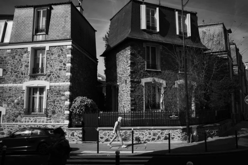 Thierry · 14/03/2024 · 3840 x 2560 Cette photographie en noir et blanc met en scène une maison en pierre de style traditionnel, probablement européenne. Le sujet principal est cette demeure à deux niveaux, avec des murs en pierre apparente et un toit pentu.La composition est symétrique, encadrée par deux bâtiments similaires. Le cadrage est serré, centré sur la maison principale. La lumière, douce et contrastée, accentue les textures et crée des ombres marquées, renforçant l’ambiance dramatique. Les couleurs dominantes, bien que monochromes, jouent sur les nuances de gris et de noir, soulignant les détails architecturaux.Le style est documentaire et esthétique, évoquant une ambiance mélancolique et intemporelle.