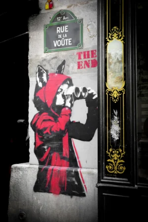 Claude · 4000 x 6000 Cette photographie montre un graffiti mural représentant un personnage en cape rouge, inspiré du style de *The Joker*, avec une tête de chat. Le sujet principal est ce personnage stylisé, situé près d’un panneau indiquant "Rue de la Voute".La composition est verticale, avec un cadrage serré sur le graffiti et une porte dorée en arrière-plan. Les couleurs dominantes sont le rouge vif, le noir et le blanc, créant un contraste marqué. L’ambiance est urbaine et artistique, renforcée par l’éclairage nocturne qui accentue les ombres et les détails. Le style est street art, dynamique et expressif.