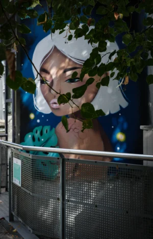 Claude · 3242 x 5063 Cette photographie montre un mural représentant une femme stylisée, les yeux fermés, avec des éléments floraux et des motifs célestes autour de sa tête. La composition est verticale, avec un cadrage serré sur le visage et les épaules.La lumière est naturelle, tamisée par des feuilles en premier plan. Les couleurs dominantes sont des bleus profonds, des verts et des touches de blanc. Le style est artistique et onirique, mêlant réalisme et éléments fantastiques. L’ambiance est paisible et mystérieuse, évoquant la sérénité et la connexion avec la nature.