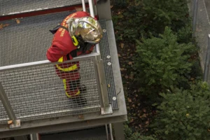 Thierry · 06/09/2025 · 3840 x 2560 Cette photographie montre un pompier, sujet principal, vêtu d'une tenue de protection rouge et jaune, équipé d’un casque et d’un harnais de sécurité. Il se trouve sur une structure métallique en hauteur, probablement un échafaudage.La composition est centrée sur le sujet, avec un cadrage serré qui met en valeur son activité. La lumière semble naturelle, bien que légèrement contrastée, créant des ombres marquées. Les couleurs dominantes sont les tons rouges, jaunes et verts (végétation en arrière-plan).Le style est documentaire et réaliste, évoquant une ambiance de travail intense et sécurisé.