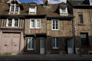 Thierry · 27/09/2025 · 3840 x 2560 Cette photographie montre une façade de maisons en pierre de style traditionnel européen, probablement du XIXe siècle. Le sujet principal est une rangée de trois bâtiments alignés, avec des fenêtres à petits carreaux et des portes en bois.La composition est symétrique et équilibrée, avec un cadrage serré sur les maisons. La lumière naturelle, douce et légèrement diffuse, met en valeur les textures de la pierre et du bois. Les couleurs dominantes sont les tons gris-beige de la pierre, contrastant avec le bois sombre des portes et des encadrements. L’ambiance est calme et rétro, évoquant un charme historique et une certaine mélancolie.