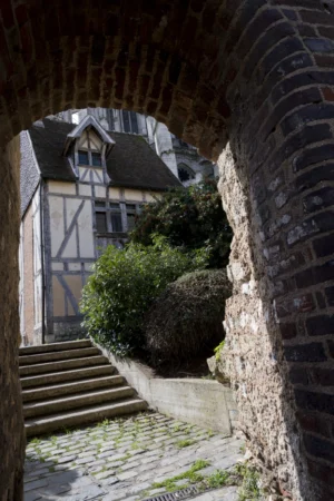 Thierry · 27/09/2025 · 2560 x 3840 Cette photographie met en scène un **maison à colombages** médiévale, sujet principal, encadrée par une **archivolte en pierre**. La composition joue sur la perspective avec un **escalier en pierre** menant vers la maison, bordé de murs anciens. Le cadrage est serré, guidant le regard vers le haut.La lumière, naturelle et douce, crée des **ombres marquées**, accentuant le relief des pierres et des poutres. Les **couleurs dominantes** sont les tons **beige, gris et verts** des murs, des pierres et de la végétation.Le style évoque un **patrimoine historique**, tandis que l’ambiance est **sereine et mystérieuse**, renforcée par les jeux de lumière et l’architecture rustique.