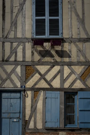Thierry · 27/09/2025 · 2560 x 3840 Cette photographie met en scène une façade de maison à colombages, sujet principal typique de l'architecture traditionnelle européenne. La composition est verticale, centrée sur les poutres en bois croisées et les fenêtres aux volets bleus fermés.Le cadrage serré accentue les détails architecturaux et les textures. La lumière naturelle, douce et légèrement dorée, souligne les contrastes entre les bois clairs et les zones d’ombre. Les couleurs dominantes sont les tons beiges des colombages, les bleus des volets et les touches de rouge et vert des menuiseries.Le style est rustique et historique, créant une ambiance chaleureuse et nostalgique.