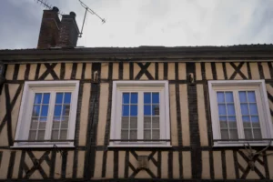 Thierry · 27/09/2025 · 3840 x 2560 Cette photographie met en scène une façade de maison à colombages, sujet principal typique de l'architecture traditionnelle européenne. La composition est symétrique, centrée sur trois fenêtres alignées, encadrées par des poutres en bois foncé.Le cadrage serré isole la partie supérieure du bâtiment, accentuant les détails architecturaux. La lumière, tamisée et légèrement grise, crée une ambiance sobre et mélancolique. Les couleurs dominantes sont les tons de bois brun et les gris clairs des murs, renforçant un style rustique et historique. L'atmosphère évoquée est à la fois calme et nostalgique.