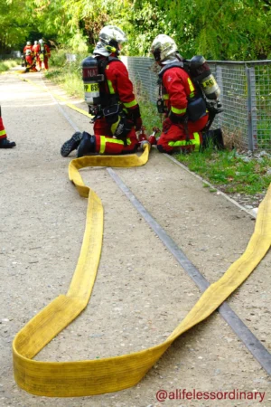 Cédric · 06/09/2025 · 3322 x 4984 Cette photographie montre deux pompiers en tenue de protection, équipés de casques et d'appareils respiratoires, assis sur un trottoir. Le sujet principal est une longue bande de sauvetage jaune déployée devant eux.La composition est dynamique, avec un cadrage serré sur les pompiers et la bande. La lumière naturelle éclaire la scène, mettant en valeur les couleurs vives. Les couleurs dominantes sont le jaune vif de la bande, le rouge et noir des tenues des pompiers, et le vert des arbres en arrière-plan.L’ambiance est professionnelle et préparatoire, évoquant une opération de secours ou un exercice. Le style est réaliste et documentaire.