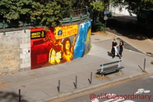 Cédric · 06/09/2025 · 5127 x 3418 Cette photographie met en scène un **mural coloré** comme sujet principal, représentant des visages stylisés aux couleurs vives. La composition inclut une **rue en pente** avec des passants, dont deux personnes en bas à droite.Le **cadrage** est aérien, offrant une vue d'ensemble. La lumière est naturelle, probablement en journée, éclairant uniformément la scène. Les **couleurs dominantes** sont vives : rouge, jaune, bleu et noir.Le style est **urbain et artistique**, mêlant street art et vie quotidienne. L’ambiance est dynamique, contrastant entre la créativité du graffiti et le cadre ordinaire de la rue.