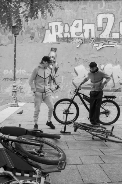 Alice · 13/09/2025 · 3270 x 4905 Cette photographie en noir et blanc montre deux hommes près de vélos. Le sujet principal semble être l’un d’eux, en train d’observer ou de discuter avec l’autre, qui est penché sur un vélo.La composition est simple et naturelle, avec un cadrage serré sur les personnages et les vélos. La lumière est douce, sans ombres marquées, créant une ambiance calme et quotidienne. Les couleurs, bien que monochromes, jouent sur les contrastes de tons clairs et foncés.Le style est documentaire, évoquant une scène de rue authentique et spontanée. L’ambiance est sobre et réaliste, renforcée par le flou des éléments en arrière-plan.