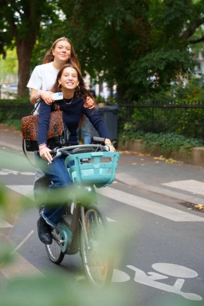 Alice · 13/09/2025 · 3637 x 5456 Cette photographie montre deux femmes sur un vélo, l’une pédalant et l’autre assise dans un panier coloré. Le sujet principal est leur complicité et leur mouvement.La composition est dynamique, avec un cadrage serré sur le vélo et les deux femmes. L’arrière-plan flou met l’accent sur elles. La lumière semble naturelle, probablement en journée, avec des ombres douces. Les couleurs dominantes sont les tons verts (végétation), bleus (panier) et neutres (vêtements).Le style est spontané et joyeux, évoquant une ambiance décontractée et amusante, typique d’un moment de partage en extérieur.
