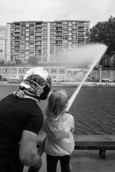 Alice · 13/09/2025 · 4463 x 6694 Cette photographie en noir et blanc montre un adulte et un enfant comme sujets principaux. L’adulte, vêtu d’un casque de football américain, tient une lance à eau dirigée vers la droite, tandis que l’enfant, debout à ses côtés, observe.La composition est dynamique, avec un cadrage serré sur les deux personnages, mettant l’accent sur leur interaction. L’arrière-plan montre un bâtiment et un plan d’eau, ajoutant de la profondeur. La lumière semble naturelle, douce, sans ombres marquées. Le style est simple et spontané, évoquant une ambiance ludique et joyeuse malgré le monochrome.