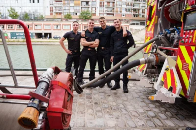 Alice · 13/09/2025 · 6315 x 4210 Cette photographie montre quatre pompiers posant devant un camion de secours. Le sujet principal est leur unité et leur équipement, notamment une grande lance à incendie.La composition est symétrique, avec les hommes alignés et le camion en arrière-plan. Le cadrage est serré, mettant en valeur les détails techniques. La lumière semble naturelle, légèrement douce, créant des ombres peu marquées.Les couleurs dominantes sont les tons sombres des tenues des pompiers, contrastant avec le rouge vif du camion et les teintes neutres du pavé. L’ambiance est professionnelle et sérieuse, évoquant la préparation et la solidarité.