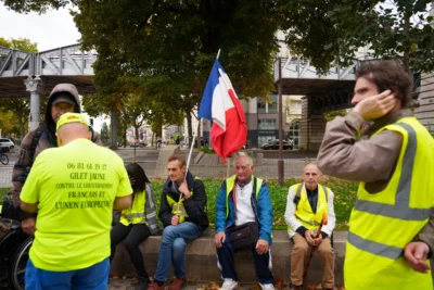 Alice · 13/09/2025 · 7008 x 4672 Cette photographie montre un groupe de manifestants assis sur des barricades. Le sujet principal est un homme tenant un drapeau français, entouré de personnes vêtues de gilets jaunes, symboles des Gilets Jaunes.La composition est dynamique, avec un cadrage serré sur les manifestants. La lumière naturelle éclaire la scène, mettant en valeur les visages et les couleurs vives. Les couleurs dominantes sont le jaune des gilets, le bleu-blanc-rouge du drapeau, et les tons neutres des vêtements.Le style est documentaire, capturant un moment authentique. L’ambiance est à la fois solennelle et combative, reflétant un esprit de protestation.