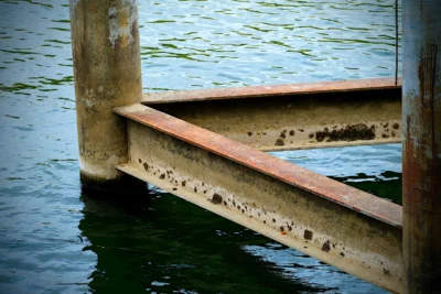 Denis · 13/09/2025 · 4898 x 3265 Cette photographie met en scène un **ponton en béton et métal rouillé** partiellement immergé dans l’eau. Le **sujet principal** est ce support érodé, soulignant son usure et son contact avec l’eau.La **composition** est épurée, avec un **cadrage serré** centré sur les éléments structurels. La **lumière**, douce et naturelle, crée des contrastes entre ombres et reflets. Les **couleurs dominantes** sont les tons gris-beige du béton, le rouge rouille du métal et les bleus-verts de l’eau.Le **style** est réaliste et documentaire, évoquant une **ambiance mélancolique et industrielle**, presque abandonnée.