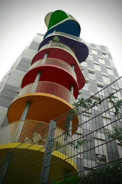 Denis · 13/09/2025 · 3265 x 4898 Cette photographie met en scène une structure architecturale colorée et moderne, probablement un escalier extérieur ou une tour d'accès. Le sujet principal est cette tour aux couleurs vives (orange, jaune, rouge, vert, bleu), disposées en cercles superposés.La composition est verticale, avec un cadrage serré qui accentue la hauteur de la structure. La lumière est naturelle, légèrement diffuse, créant des contrastes doux. Les couleurs dominantes sont vives et saturées, apportant une touche joyeuse et dynamique. Le style est contemporain et artistique, évoquant une ambiance ludique et innovante dans un environnement urbain.