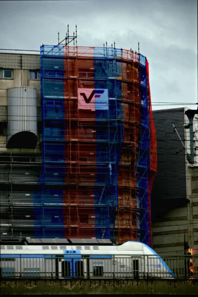 Denis · 13/09/2025 · 3265 x 4898 Cette photographie montre un immeuble en cours de rénovation, recouvert de filets de protection colorés (bleu, rouge et transparent). Le sujet principal est le bâtiment avec un logo "V" visible. La composition met en avant la verticalité et la structure architecturale.Le cadrage est serré, centré sur l'immeuble. La lumière est diffuse, probablement par temps nuageux, créant une ambiance sobre. Les couleurs dominantes sont les teintes bleues et rouges des filets, contrastant avec le gris du ciel et des bâtiments environnants. Le style est documentaire et réaliste, évoquant une ambiance urbaine et industrielle.