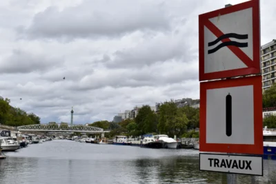 Deniz · 13/09/2025 · 6000 x 4000 Cette photographie montre un panneau de signalisation indiquant des travaux ("TRAVAUX") avec des symboles de restrictions (accès interdit aux piétons et véhicules).La composition met en avant le panneau en premier plan, tandis que l’arrière-plan révèle un canal avec des bateaux et des bâtiments. Le cadrage est serré sur le panneau, mais inclut une vue dégagée sur le paysage urbain.La lumière est douce, légèrement voilée par des nuages. Les couleurs dominantes sont les tons gris-bleu du ciel et de l’eau, contrastant avec le rouge vif du panneau. L’ambiance est calme et urbaine, évoquant une journée ordinaire en ville.