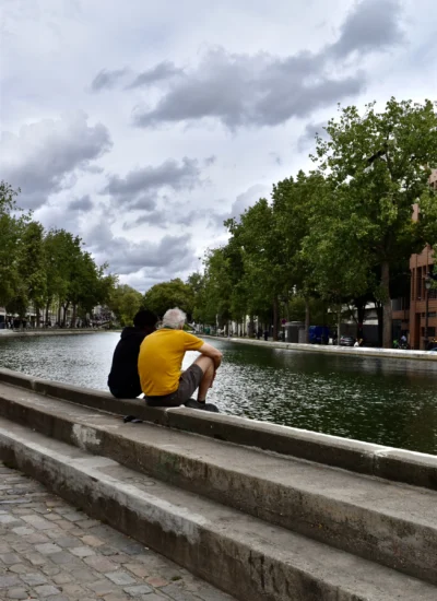 Deniz · 13/09/2025 · 4000 x 5494 Cette photographie montre une personne assise en tailleur sur une margelle de canal, vêtue d’un pull jaune. Le sujet principal est flou, suggérant un mouvement ou une intention artistique.La composition repose sur une perspective longitudinale, avec le canal bordé d’arbres en arrière-plan. Le cadrage inclut une partie du ciel nuageux et des bâtiments discrets.La lumière est naturelle, douce, avec des ombres peu marquées. Les couleurs dominantes sont les verts des arbres, le gris du ciel et du béton, et le jaune vif du vêtement.Le style est sobre et réaliste, évoquant une ambiance calme et contemplative, presque mélancolique.