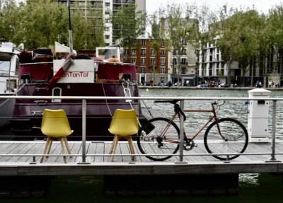 Deniz · 13/09/2025 · 5579 x 3998 Cette photographie met en scène un vélo noir attaché à une rambarde en métal, avec deux chaises jaunes à côté. Le sujet principal est ce vélo, symbolisant peut-être une pause ou une attente.La composition est simple et équilibrée, avec un cadrage serré sur le premier plan. En arrière-plan, on aperçoit un canal, des bâtiments et des arbres, créant une profondeur.La lumière semble naturelle, légèrement tamisée, donnant une ambiance calme et paisible. Les couleurs dominantes sont les tons gris (bâtiments, eau), jaune (chaises) et noir (vélo), renforçant une atmosphère urbaine et sereine. Le style est réaliste et documentaire.