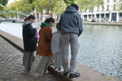 Evelyne · 3840 x 2560 Cette photographie montre un groupe de quatre personnes près d’un canal urbain. Le sujet principal semble être les interactions entre eux, avec un homme en sweat-shirt gris et trois autres personnes, dont une enfant.La composition est dynamique, avec un cadrage serré sur les individus. La lumière est naturelle, probablement en journée, avec des ombres douces. Les couleurs dominantes sont les tons neutres (gris, beige) et quelques touches de couleurs vives comme le rouge de la veste de l’enfant.Le style est documentaire et spontané, capturant un moment de vie quotidienne. L’ambiance est calme et urbaine, évoquant une scène de détente en ville.