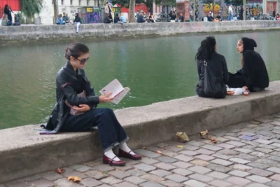 Evelyne · 3840 x 2560 Cette photographie montre trois jeunes femmes assises sur une berge en pierre près d’un canal. Le sujet principal est la femme à gauche, lisant un livre avec des lunettes de soleil.La composition est équilibrée, avec un cadrage serré sur les personnes et l’eau en arrière-plan. La lumière semble naturelle, douce, et légèrement tamisée, créant des ombres douces. Les couleurs dominantes sont les tons neutres (gris, bleu du canal) et les vêtements sombres (noir, bleu marine).Le style est réaliste et documentaire, capturant une scène du quotidien. L’ambiance est calme et studieuse, évoquant une pause détendue en ville.