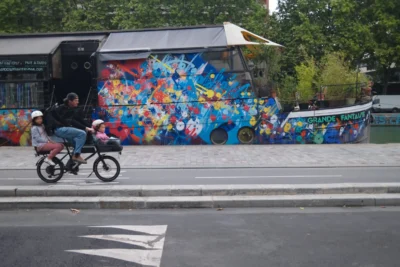Evelyne · 3840 x 2560 Cette photographie met en scène une femme et un enfant à vélo, passant devant un bus décoré de fresques murales colorées. Le sujet principal est le mouvement et la vie urbaine, avec une touche artistique.La composition joue sur la juxtaposition entre le dynamisme des cyclistes et la statique des couleurs vives du bus. Le cadrage est serré sur les personnages, avec un arrière-plan artistique. La lumière semble naturelle, probablement en journée.Les couleurs dominantes sont vives et contrastées : bleus, roses, jaunes et verts. Le style est urbain et artistique, créant une ambiance joyeuse et vivante.