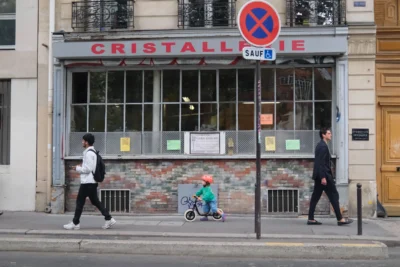 Evelyne · 3840 x 2560 Cette photographie montre une rue urbaine devant un magasin nommé **« Cristallerie »**. Le sujet principal est une scène de vie quotidienne : trois personnes (dont un enfant sur un trottinette) traversent ou marchent près d’un passage piéton.La composition est dynamique, avec un cadrage serré sur le magasin et ses vitrines. La lumière est naturelle, légèrement tamisée, créant des ombres douces. Les couleurs dominantes sont les tons gris (bâtiments), verts (enseignes) et bleus (ciel). L’ambiance est calme et réaliste, reflétant une ville animée mais paisible. Le style est documentaire et spontané.