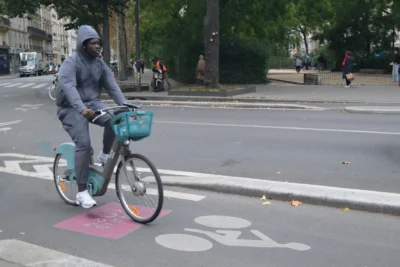 Evelyne · 3840 x 2560 Cette photographie montre un cycliste vêtu d'une combinaison grise et d'un masque, roulant sur un vélo bleu avec panier. Le sujet principal est bien mis en valeur par le cadrage serré et la position centrale.La composition utilise une perspective légèrement basse, avec des éléments urbains en arrière-plan (arbres, trottoir, passants). La lumière semble naturelle, un peu diffuse, typique d’un jour nuageux. Les couleurs dominantes sont les tons gris et bleus, renforçant une ambiance sobre et moderne. L’atmosphère est calme et quotidienne, reflétant une scène urbaine banale.