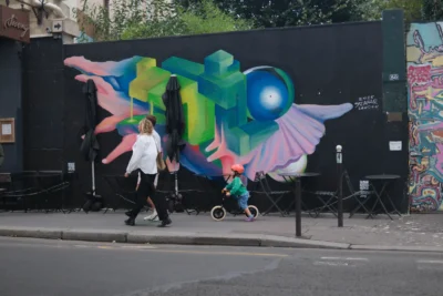 Evelyne · 3840 x 2560 Cette photographie met en scène un **mural coloré** représentant une créature fantastique aux ailes irisées, dominant la composition. Au premier plan, une femme en blanc pousse un enfant sur un vélo, ajoutant une touche de vie urbaine.Le **cadrage** est serré sur le mur, avec une perspective légèrement basse. La **lumière** naturelle éclaire la scène, mettant en valeur les **couleurs vives** (bleu électrique, vert, rose) du graffiti. Le style est **urbain et artistique**, dynamique, créant une ambiance à la fois joyeuse et onirique, mêlant art de rue et quotidien.