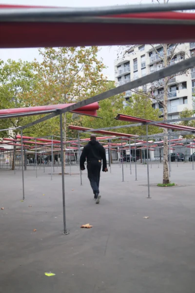 Evelyne · 2560 x 3840 Cette photographie montre un homme marchant sous une structure de tentes fermées, probablement un marché ou une foire non ouverte. Le sujet principal est cet individu, vu de dos, en mouvement.La composition utilise des lignes horizontales et verticales pour guider le regard. Le cadrage est serré, avec un premier plan légèrement flou et un arrière-plan plus net. La lumière est naturelle, tamisée, créant des ombres douces. Les couleurs dominantes sont les tons gris et marron, avec des touches de rouge sur les tentes.L’ambiance est calme et un peu mélancolique, évoquant un lieu temporairement inactif.