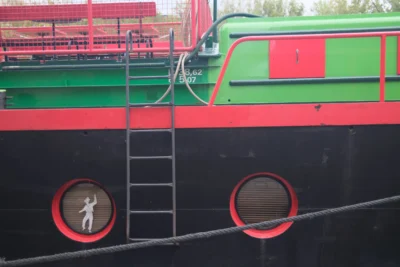 Evelyne · 3840 x 2560 Cette photographie met en scène un bateau aux couleurs vives. Le sujet principal est une échelle de corde avec une silhouette humaine en train de monter, reflétée dans les hublots circulaires rouges et noirs.La composition est centrée sur les détails techniques du bateau. Le cadrage serré isole les éléments, créant un effet visuel dynamique. Les couleurs dominantes sont le noir, le rouge et le vert, apportant un contraste marqué. La lumière semble naturelle, bien que légèrement tamisée, renforçant l’ambiance industrielle et maritime. Le style est réaliste et documentaire, évoquant une atmosphère à la fois robuste et mystérieuse.