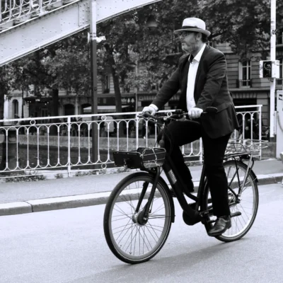 Fabienne · 13/09/2025 · 3840 x 3840 Cette photographie en noir et blanc met en scène un homme en costume élégant, chapeau et cravate, comme sujet principal. Il est assis sur un vélo classique, en mouvement.La composition est simple et équilibrée, avec une perspective légèrement basse. Le cadrage isole le sujet sur la route, encadré par une rambarde et des arbres en arrière-plan.La lumière semble naturelle, douce, créant des contrastes modérés. Les couleurs dominantes sont les nuances de gris typiques du noir et blanc.Le style évoque une ambiance rétro, intemporelle et sophistiquée, rappelant les époques passées. L'ambiance est calme et élégante.