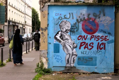 Fabienne · 3840 x 2560 Cette photographie montre un mur tagué avec des graffitis en français et en arabe, dont la phrase *« On pisse pas ici »*. Le sujet principal est le graffiti, avec une silhouette stylisée et des symboles politiques.La composition inclut deux personnes en arrière-plan, floutées, marchant sur un trottoir urbain. Le cadrage est serré sur le mur, avec une perspective légèrement basse. La lumière est naturelle, diffuse, typique d’un jour couvert. Les couleurs dominantes sont les tons bleus, blancs et gris, renforçant une ambiance urbaine et légèrement mélancolique, mêlant protestation et vie quotidienne.