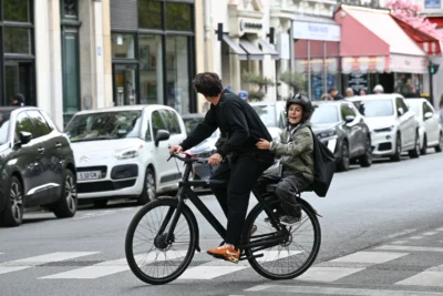 Jean-Marc · 13/09/2025 · 3840 x 2560 Cette photographie met en scène deux personnes à vélo en ville. Le sujet principal est le cycliste en premier plan, vêtu d’un manteau sombre, avec un passager assis derrière lui.La composition montre une rue urbaine avec des voitures en arrière-plan et des bâtiments. Le cadrage est serré sur les cyclistes, capturant leur mouvement.La lumière semble naturelle, probablement en journée, avec des ombres douces. Les couleurs dominantes sont les tons gris et neutres, adoucis par des touches de vert et de rouge (voiture et enseigne).Le style est documentaire et réaliste, évoquant une ambiance dynamique et quotidienne en milieu urbain.
