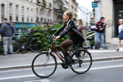 Jean-Marc · 13/09/2025 · 3840 x 2560 Cette photographie met en scène une jeune femme comme sujet principal, pédalant sur un vélo en ville. La composition est dynamique, avec un cadrage serré sur elle, légèrement flou en arrière-plan, créant un effet de mouvement.La lumière semble naturelle, probablement en journée, avec des ombres douces. Les couleurs dominantes sont neutres (gris de la ville, noir de ses vêtements) rehaussées par des touches de rouge (vélo) et de vert (végétation).Le style est documentaire et spontané, capturant une scène urbaine authentique. L’ambiance est vivante et moderne, reflétant le quotidien citadin.