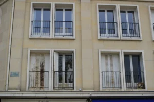 Marie-Odile · 1600 x 1066 Cette photographie montre la façade d’un immeuble résidentiel de style classique européen. Le sujet principal est une série de fenêtres et balcons en fer forgé, alignés verticalement.La composition est symétrique, avec des lignes nettes et répétitives. Le cadrage est serré, mettant en valeur les détails architecturaux. La lumière est naturelle, douce et diffuse, sans ombres marquées. Les couleurs dominantes sont des tons neutres : beige clair pour les murs, gris pour les balcons et blanc pour les fenêtres.L’ambiance est sobre et élégante, évoquant un quartier urbain paisible et traditionnel.