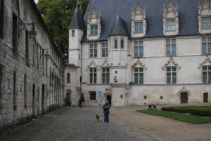Marie-Odile · 1600 x 1066 Cette photographie montre un bâtiment historique aux allures médiévales, probablement un château ou un manoir. Le sujet principal est l’architecture avec ses tourelles, ses fenêtres à meneaux et ses murs en pierre.La composition est symétrique, avec une cour centrale pavée et des bâtiments alignés. Le cadrage inclut une personne en bas à droite pour donner l’échelle. La lumière est naturelle et douce, créant des ombres légères. Les couleurs dominantes sont les tons gris-beige des pierres et les verts des végétations.L’ambiance est calme et historique, évoquant un lieu chargé d’histoire.
