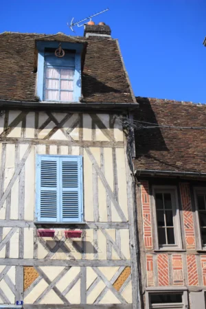 Marie-Odile · 1066 x 1600 Cette photographie montre une façade de maison à colombages, typique de l'architecture traditionnelle européenne.Le sujet principal est cette structure en bois rempli de torchis, avec des fenêtres à volets bleus fermés. La composition met en valeur les lignes diagonales des poutres et la symétrie des ouvertures. Le cadrage est serré, centré sur la partie supérieure de la maison.La lumière est naturelle et vive, créant des ombres nettes. Les couleurs dominantes sont les tons beiges et bruns du bois, contrastés par les bleus des volets et les touches de rouge et jaune des encadrements. L’ambiance est rustique et chaleureuse, évoquant un charme ancien et pittoresque.