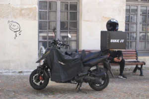 Marie-Odile · 1600 x 1066 Cette photographie met en scène un **scooter couvert** comme sujet principal, garé devant une façade urbaine. La **composition** associe le véhicule à un banc et une porte close, avec un graffiti discret sur le mur.Le **cadrage** est serré, centré sur le scooter et son environnement immédiat. La **lumière** semble naturelle, tamisée, créant des ombres douces. Les **couleurs dominantes** sont neutres : gris, noir et beige, avec des touches de bleu sur le scooter.Le style est **documentaire et urbain**, évoquant une ambiance **calme et quotidienne**, presque mélancolique.