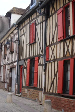 Marie-Odile · 1066 x 1600 Cette photographie montre une rue bordée de maisons à colombages, typiques de l'architecture médiévale française. Le sujet principal est ces maisons en bois, avec des poutres apparentes et des encadrements de fenêtres rouges.La composition est symétrique et linéaire, avec un cadrage serré sur les façades. La lumière naturelle, douce et légèrement tamisée, crée des ombres douces. Les couleurs dominantes sont les tons beiges des bois, contrastés par le rouge des fenêtres et les touches de vert et de gris.L’ambiance est chaleureuse et pittoresque, évoquant un charme historique et rustique.