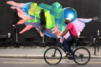 Fabienne · 3840 x 2559 Cette photographie met en scène un cycliste comme sujet principal, vêtu d’un t-shirt rouge et d’un masque, en mouvement devant un mur. La composition joue sur le contraste entre le sujet dynamique et le fond statique.Le cadrage est serré sur le cycliste, avec une partie du mur coloré en arrière-plan. La lumière semble naturelle, bien répartie, sans ombre marquée. Les couleurs dominantes sont vives : bleu, vert, rouge et jaune sur le mural, créant un style urbain et artistique. L’ambiance est à la fois moderne et énergique, mêlant mouvement et créativité.