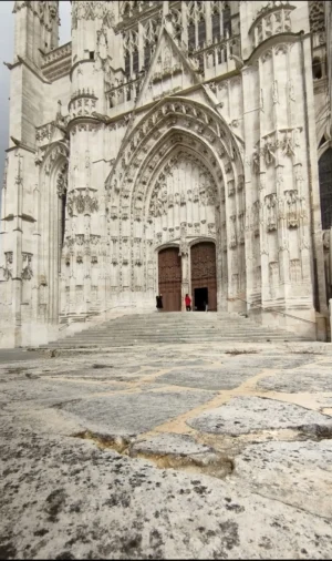 Muriel · 1080 x 1822 Cette photographie montre une façade gothique d'une cathédrale. Le sujet principal est l'entrée monumentale, avec ses arcs brisés, ses sculptures et ses portes en bois.La composition est symétrique, centrée sur le portail. Le cadrage est serré, mettant en valeur les détails architecturaux. La lumière, naturelle et douce, accentue les textures et les ombres des pierres.Les couleurs dominantes sont les tons gris-beige des pierres, avec des touches de vert et de rouge sur les portes. Le style est architectural gothique, évoquant une ambiance historique, solennelle et presque mystique.