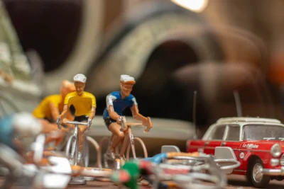 Hector · 13/09/2025 · 3840 x 2560 Cette photographie met en scène des **miniatures de cyclistes** comme sujet principal, placées sur un décor de maquettes urbaines. La **composition** montre un cadrage serré, centré sur les personnages en mouvement, avec des éléments flous en arrière-plan (voitures, bâtiments).La **lumière** semble naturelle et douce, créant des ombres légères. Les **couleurs dominantes** sont vives : jaune, bleu et rouge des vêtements, contrastant avec les tons neutres du décor.Le **style** évoque une ambiance miniature réaliste, évoquant un **diorama** ou une scène de jeu. L’**ambiance** est dynamique, presque cinématographique, avec une touche nostalgique et ludique.