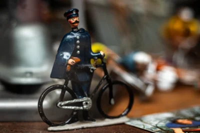Hector · 13/09/2025 · 3840 x 2560 Cette photographie met en scène une figurine de policier miniature en uniforme du XIXe siècle, assis sur un vélo. Le sujet principal est net et centré, avec un cadrage serré qui isole la figurine.L'arrière-plan est flou, créant un effet de profondeur. La lumière douce et naturelle accentue les détails de la figurine. Les couleurs dominantes sont les tons sombres (bleu, noir) de l'uniforme, contrastant avec le métal argenté du vélo.Le style évoque une ambiance nostalgique et miniaturiste, renforcée par l'aspect vintage et le flou artistique de l'arrière-plan.