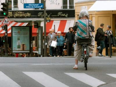 Seila · 13/09/2025 · 3840 x 2880 Cette photographie capture une personne à vélo comme sujet principal, traversant un passage piéton en ville. La composition est dynamique, avec un cadrage serré sur le cycliste, mettant en valeur son mouvement. L’arrière-plan montre une rue animée avec des passants et des enseignes.La lumière est naturelle, légèrement tamisée, créant des ombres douces. Les couleurs dominantes sont les tons neutres (gris, beige) avec des touches de rouge et bleu sur les enseignes et vêtements. Le style est documentaire et urbain, évoquant une ambiance quotidienne et vivante dans une ville européenne.