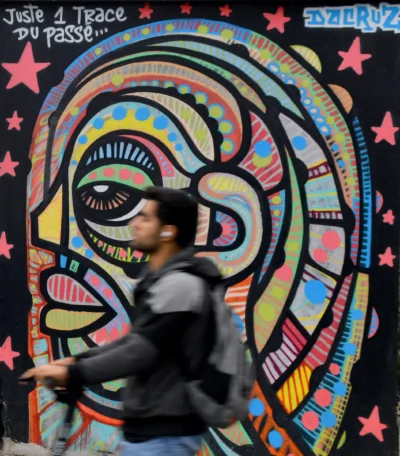 Vincent · 13/09/2025 · 3370 x 3840 Cette photographie montre une personne marchant devant un mural coloré. Le sujet principal est ce graffiti stylisé, représentant un visage aux traits psychédéliques et aux couleurs vives. La composition joue sur le contraste entre le mouvement du passant et l'immobilité de l'œuvre.Le cadrage est serré sur le mural, avec une lumière naturelle qui éclaire la scène. Les couleurs dominantes sont vives et contrastées : bleus, roses, jaunes et verts. Le style est artistique et urbain, évoquant une ambiance dynamique et créative, typique des scènes street art. L'atmosphère est à la fois énergique et festive.