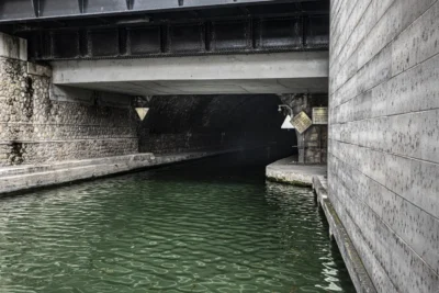 Benoit · 13/09/2025 · 1500 x 1000 Cette photographie montre un **passage souterrain sous un pont**, avec un **coulis d’eau verte** en contrebas. Le **sujet principal** est l’architecture industrielle du pont en béton, encadré par des murs de soutènement.La **composition** est symétrique, avec un **cadrage serré** mettant en valeur les structures et l’eau. La **lumière**, naturelle mais tamisée, crée des ombres douces. Les **couleurs dominantes** sont les tons gris du béton et le vert foncé de l’eau.Le style est **réaliste et sobre**, évoquant une **ambiance à la fois urbaine et mystérieuse**, presque mélancolique.