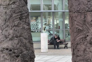 Laurence · 27/09/2025 · 2805 x 1889 Cette photographie montre un homme assis sur un banc entre deux piliers massifs et rugueux. Le sujet principal est cet homme, vêtu d’un manteau sombre, semblant se reposer ou réfléchir.La composition utilise un cadrage serré, encadrant le personnage entre les piliers, créant un effet de tunnel. La lumière est naturelle, douce mais contrastée, avec des ombres marquées. Les couleurs dominantes sont les tons gris et beiges des piliers, contrastant avec le manteau sombre de l’homme et les teintes claires du bâtiment en arrière-plan.Le style est réaliste et sobre, évoquant une ambiance calme et introspective, presque urbaine et mélancolique.