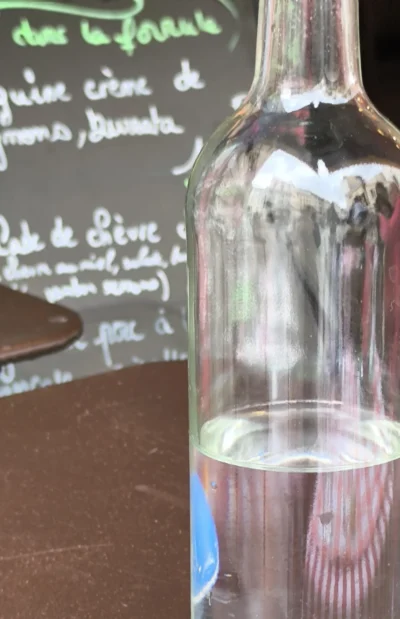 Laurence · 13/09/2025 · 2348 x 3631 Cette photographie montre une bouteille en verre remplie d’un liquide trouble, probablement du lait fermenté ou une boisson artisanale, posée devant une ardoise.Le cadrage est serré sur la bouteille, avec un arrière-plan flou où l’on distingue des inscriptions manuscrites en français. La lumière naturelle éclaire la scène, créant des reflets sur le verre. Les couleurs dominantes sont les tons froids (bleutés, gris) et le bois sombre de la table.L’ambiance est artisanale et rustique, évoquant un contexte traditionnel ou local, peut-être un marché ou une ferme. Le style est simple et documentaire.