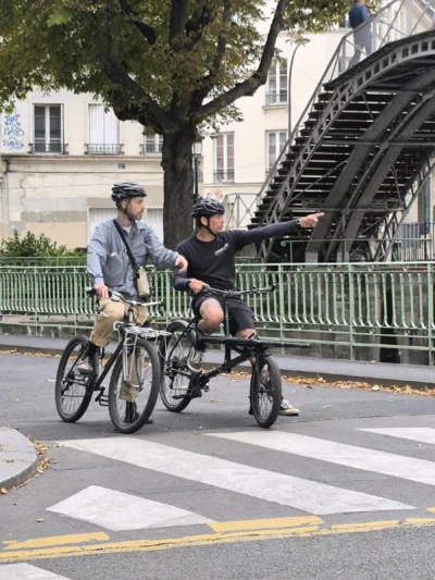Laurence · 13/09/2025 · 3060 x 4080 Cette photographie montre deux personnes à vélo, dont l’une semble guider l’autre sur un vélo tandem. Le sujet principal est ce duo en mouvement.La composition met en valeur la rue urbaine avec une perspective légèrement basse, encadrée par une grille et des escaliers en arrière-plan. Le cadrage est serré, capturant les détails des vélos et des vêtements des cyclistes.La lumière est naturelle et semble douce, typique d’une journée ensoleillée. Les couleurs dominantes sont les tons neutres (gris des vêtements, métal des vélos) et les verts des arbres, créant une ambiance à la fois dynamique et paisible, typique d’une balade urbaine.