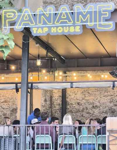 Laurence · 13/09/2025 · 2670 x 3405 Cette photographie montre l'extérieur d'un établissement nommé **"Paname Tap House"**. Le sujet principal est l'entrée du bar, avec son enseigne lumineuse et son ambiance conviviale.La composition met en avant des clients assis à des tables sous une structure ouverte. Le cadrage inclut une vue partielle de l'intérieur, avec des néons et des éléments industriels. La lumière est douce et naturelle, avec des éclairages artificiels chauds. Les couleurs dominantes sont des tons neutres (gris, beige) rehaussés par des touches de bleu et de vert. Le style est moderne et décontracté, évoquant une ambiance chaleureuse et détendue.